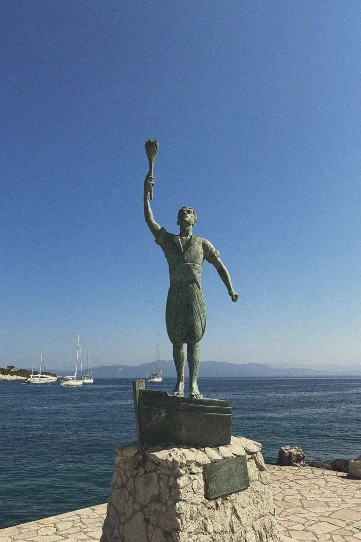 Statue of a figure raising a torch, symbolizing guidance, standing tall against the clear blue sky, with the sea and sailing boats in the background, representing Paxos's maritime heritage.